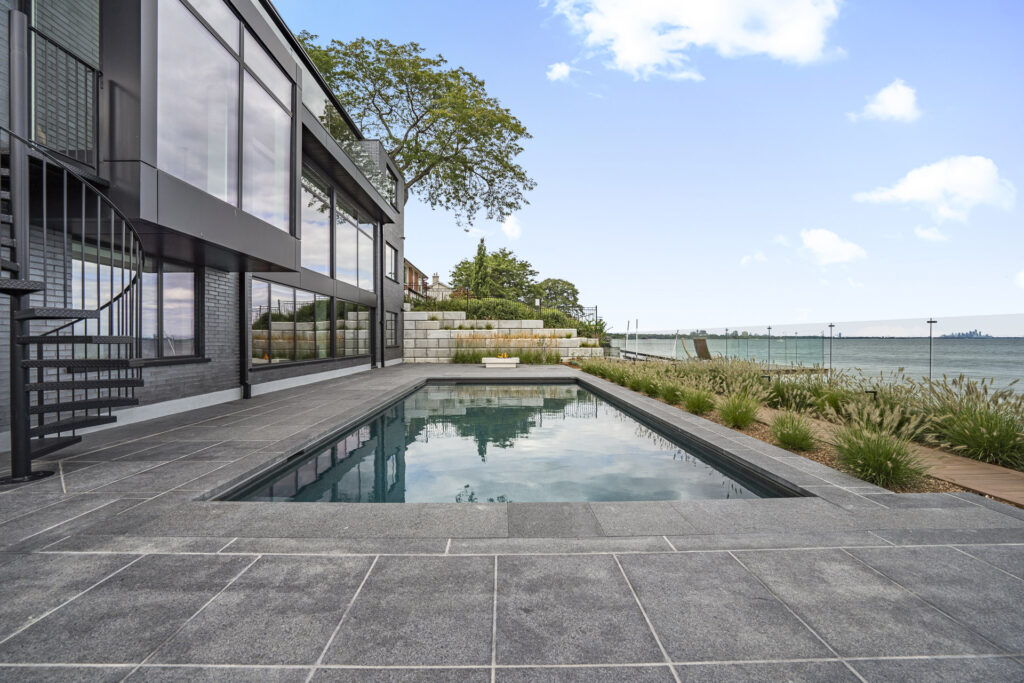 Modern lakeside house with large glass windows, a spiral staircase, and a rectangular swimming pool surrounded by gray stone tiles. Grassy landscaping and a deck overlook the water under a partly cloudy sky.